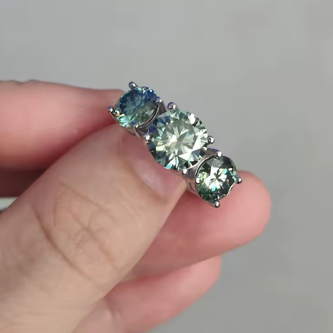 Three-stone ring with green gemstones held between fingers against a neutral background
