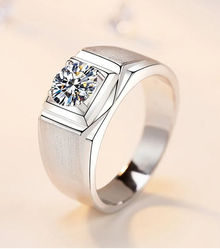 A silver-toned men's ring featuring a square bezel-set clear gemstone on a polished band, shown on a soft beige background.
