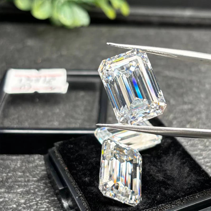 Two emerald-cut diamonds held by tweezers on a dark surface with greenery in the background.